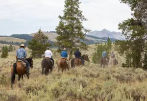 Six riders seen from behind ride single file through sagebrush and pines toward a distant rocky mountain peak