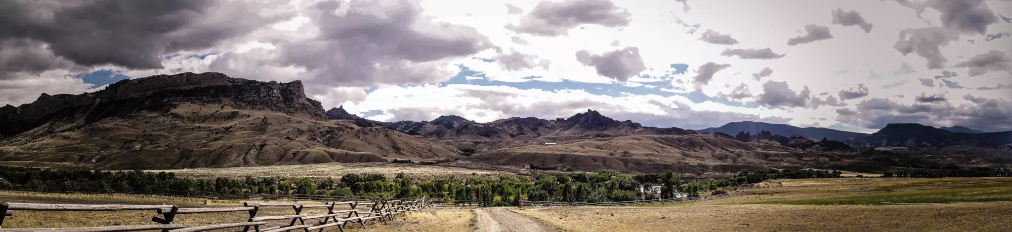 Craggy mountains and rangeland under dramatic clouds, with a split-rail fence and a dirt road in front
