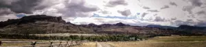 Craggy mountains and rangeland under dramatic clouds, with a split-rail fence and a dirt road in front