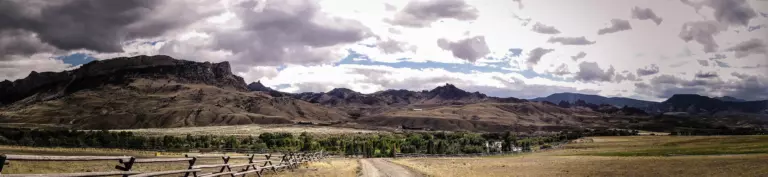 Craggy mountains and rangeland under dramatic clouds, with a split-rail fence and a dirt road in front
