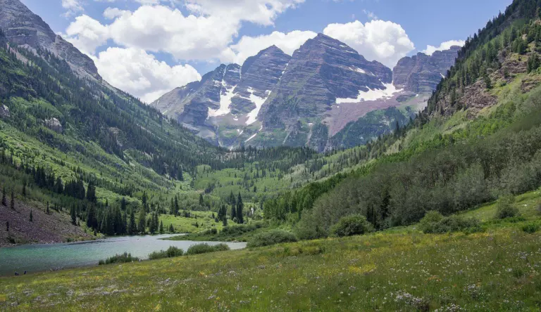 Snow-streaked Maroon Bells peaks above a green forested valley, with Maroon Lake and a wildflower meadow in the foreground under summer clouds