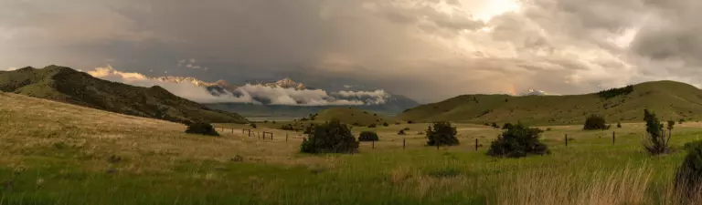 Rolling green hills and fenced pasture under storm clouds, with snowy peaks in mist beyond