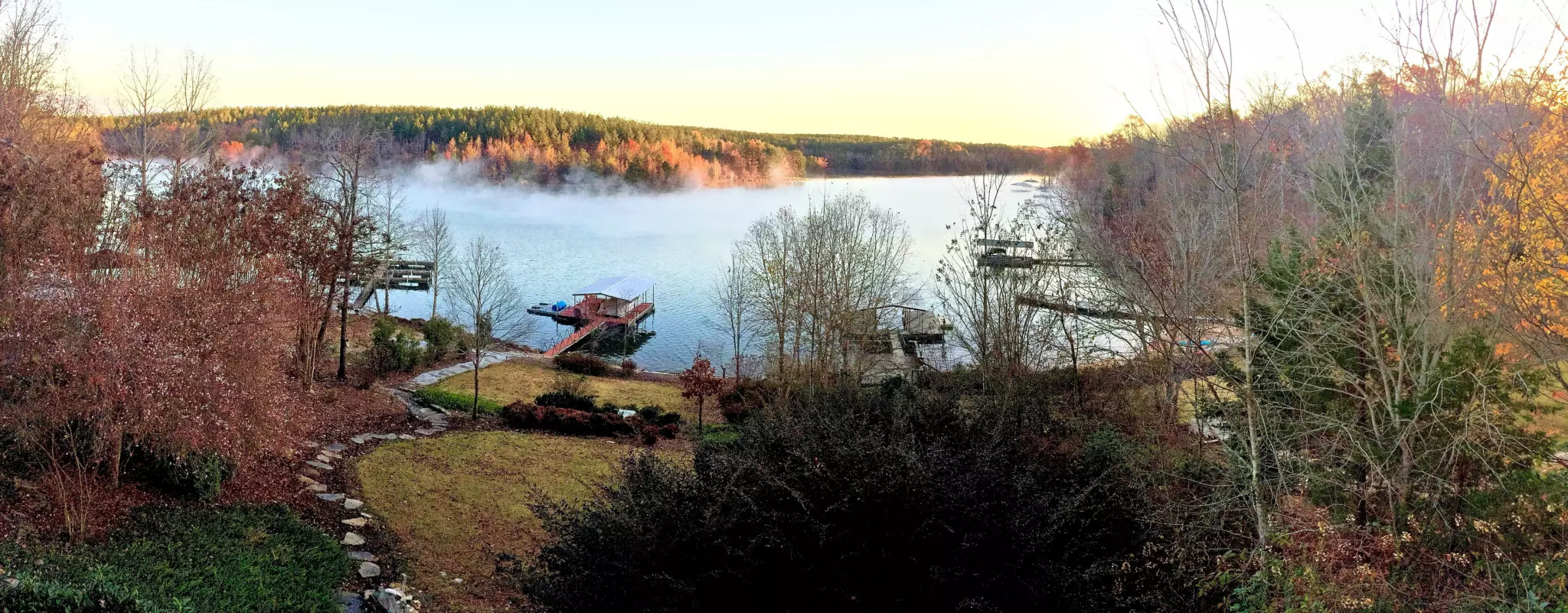 Morning mist drifting over Lake Keowee below wooded hills in autumn colour, with a boathouse, boat docks and a sloping lakeside lawn in the foreground