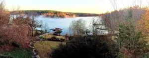Morning mist drifting over Lake Keowee below wooded hills in autumn colour, with a boathouse, boat docks and a sloping lakeside lawn in the foreground