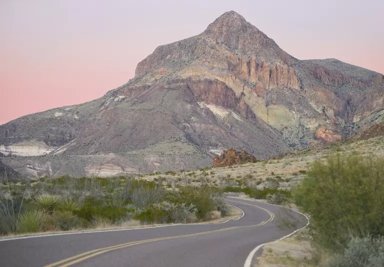 Rugged desert peak in soft dusk light under a pink sky, above yucca, green desert scrub and a winding paved road