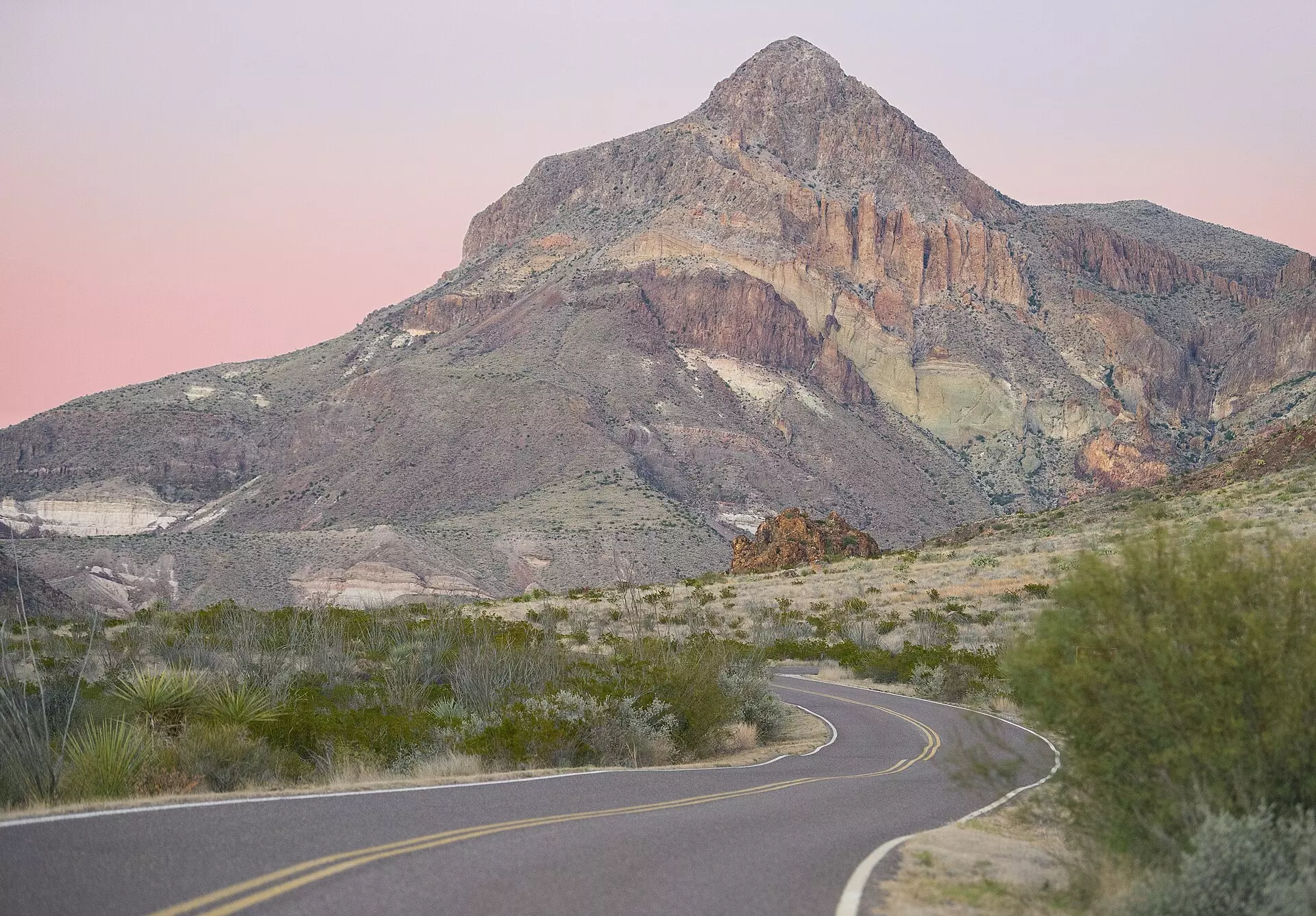 Rugged desert peak in soft dusk light under a pink sky, above yucca, green desert scrub and a winding paved road