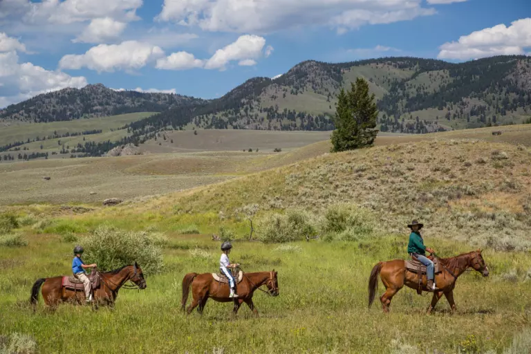 Two young riders in helmets and a wrangler ride single file through a green meadow below forested hills