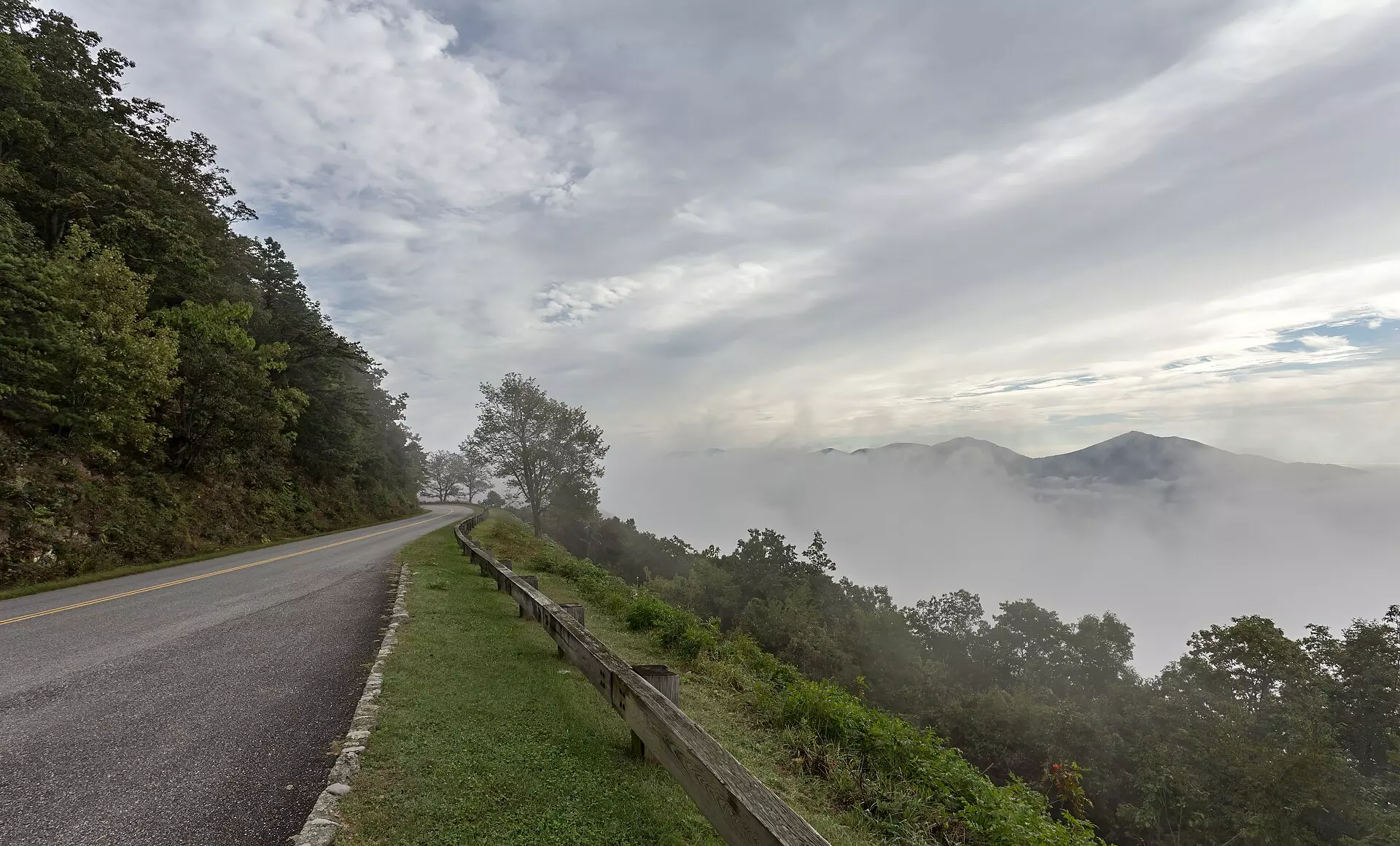 Blue Ridge Parkway curving along a wooded ridge beside a wooden guardrail, with fog filling the valley below distant mountain peaks under a pale cloudy sky